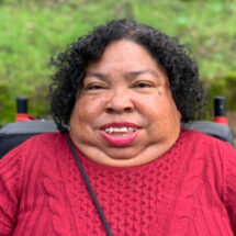 Photo of a Filipino/Black woman with short dark curly hair, wearing a knit red sweater, with a small part of the back of her wheelchair visible, in front of a background of leaves.