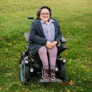 Sandy Ho is an Asian American woman sitting in a power wheelchair. She has round glasses and is wearing a gray sweater over a blue striped collar shirt, and red checkered pants with maroon shoes. She is sitting in a field of grass.