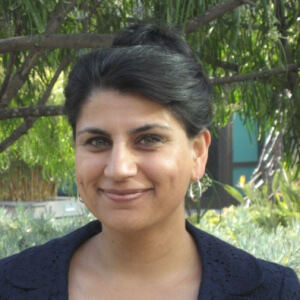Headshot of Mona Jhawar, a brown, Indian American female with hazel eyes, long dark hair pulled back into a bun, small silver hoop earrings, smiling. She is wearing a navy-blue jacket with a green blouse standing in a courtyard of green trees and foliage.