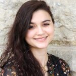 Photo of Anjali taken outdoors against a brick wall. She is wearing a floral top and gold necklace and has dark brown wavy hair.