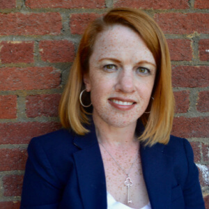 A white woman with red hair wearing a navy blazer and white top, against a brick wall.