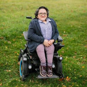 Sandy Ho is an Asian American woman sitting in a power wheelchair. She has round glasses and is wearing a gray sweater over a blue striped collar shirt, and red checkered pants with maroon shoes. She is sitting in a field of grass.