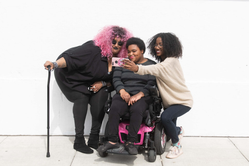 A zoomed out full body shot showing three Black and disabled friends (a non-binary person with a cane and tangle stim toy, a non-binary person sitting in a power wheelchair, and an invisibly disabled femme) smiling and taking a cell phone selfie together. All are outdoors in front of a white wall.