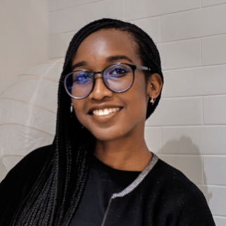 Tolu is a Black woman with long, dark braids and large, black glasses and wearing a black sweater, t-shirt and jeans. She is smiling and standing in front of a white background decorated with white paper lanterns.