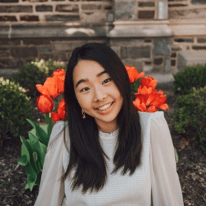 Jennifer is a Korean-American woman with medium-length black hair, wearing a white blouse. She is smiling against a background of red tulips and green bushes. 