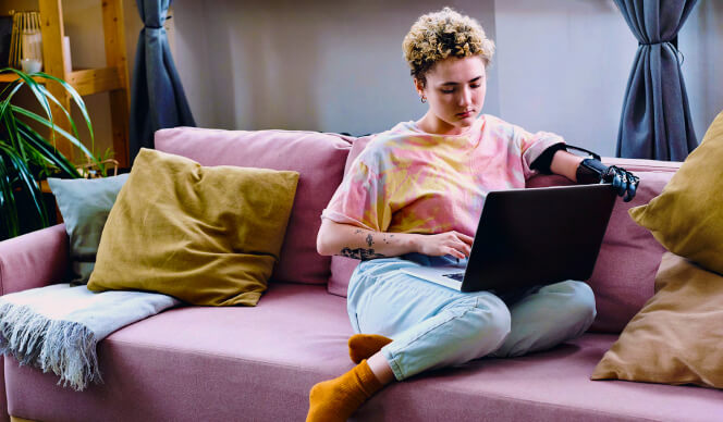 Woman with a prosthetic arm sitting on a pink sofa looking at her laptop.