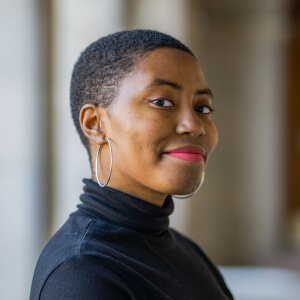 Olivia Williams, a young Black woman with low cut coily black hair, wearing a black turtleneck, silver hoop earrings, and red lipstick. She is smiling softly at the camera.