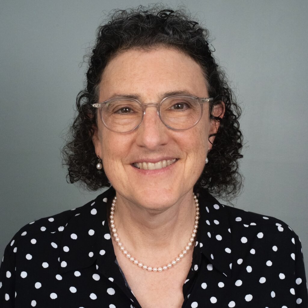 Emily Harris, a white woman with short dark curly hair wearing glasses and a black-and-white polka dot top, standing in front of a gray background.