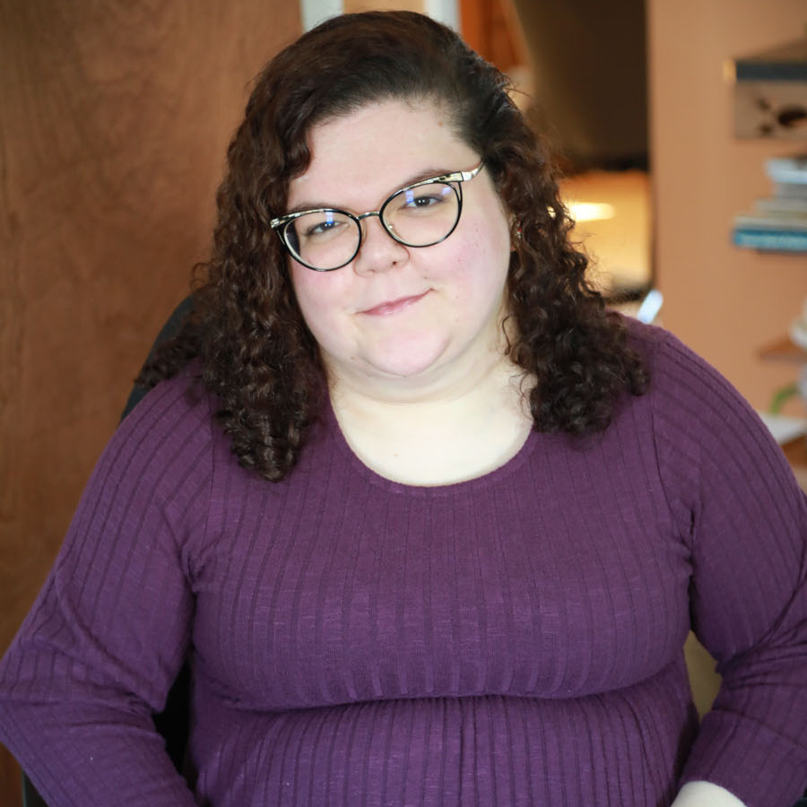 Headshot of Emily Ladau, a white woman with brown curly shoulder length hair wearing glasses and a purple sweater.