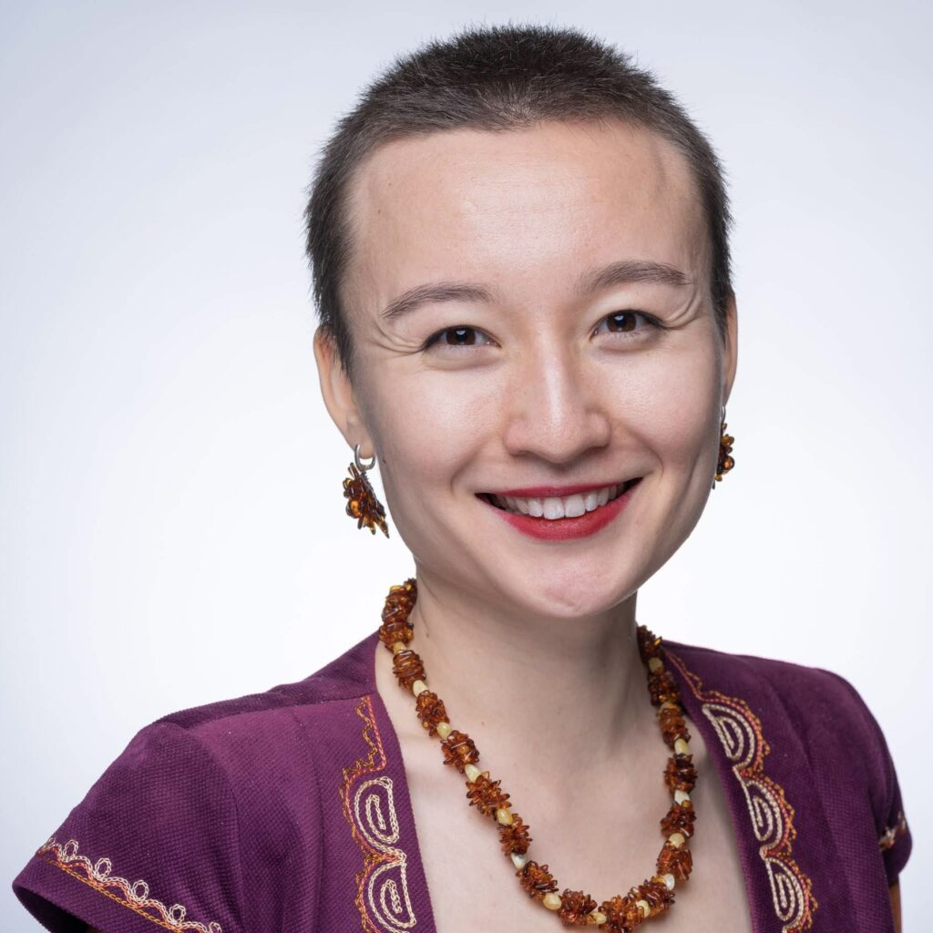 This headshot portrays an Asian woman with short hair, wearing a purple vest, a warm smile, and accessorized with an amber necklace and earrings.