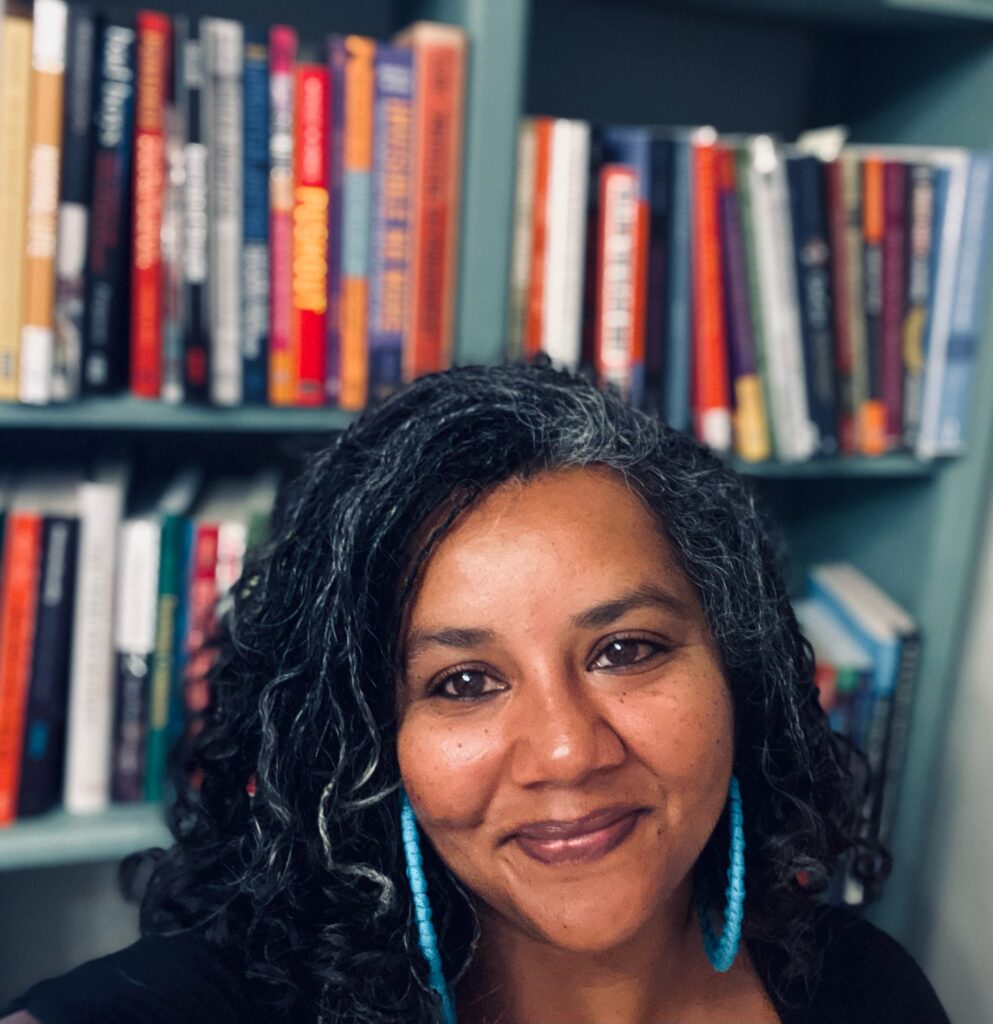 Headshot of Subini Annamma, a Black Asian woman wearing turquoise earrings, in front of a full, colorful bookshelf.