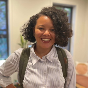 Jalyn is a Black, Japanese person with short curly hair. They stand indoors smiling while wearing a white button-up shirt with black stripes and an olive green backpack. In the background, there are windows, plants, and modern pink furniture.