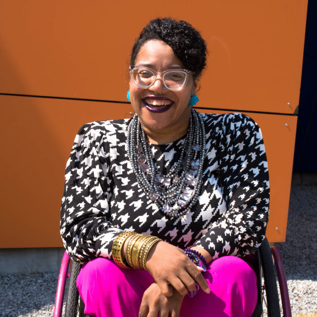 Luticha Doucette is honey skinned person sitting in a hot pink and black wheelchair with their hair braided into a swoop across their forehead. They are wearing a black and white houndstooth long sleeve shirt with fire hot pink pants, large clear glasses, Aqua colored earrings that look like rock candy, and a silver multistrand necklace.