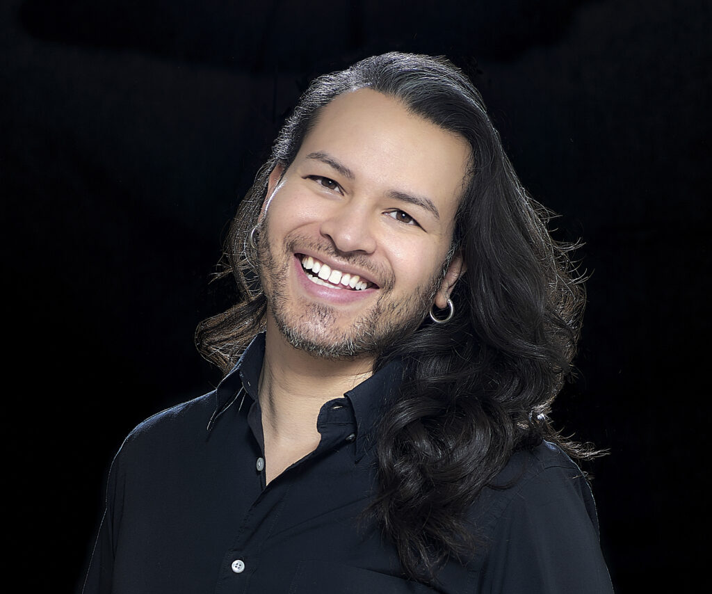 A headshot of Gabriel San Emeterio, a Mexican gender expansive person, smiling at the camera against a black background. They are wearing a dark blue button down and small hoop earrings.