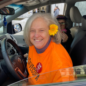 Julie Farrar, a white womam with gray short hair, wearing an orange t-shirt and a yellow flower in her hair. She is sitting in the front seat of a car.
