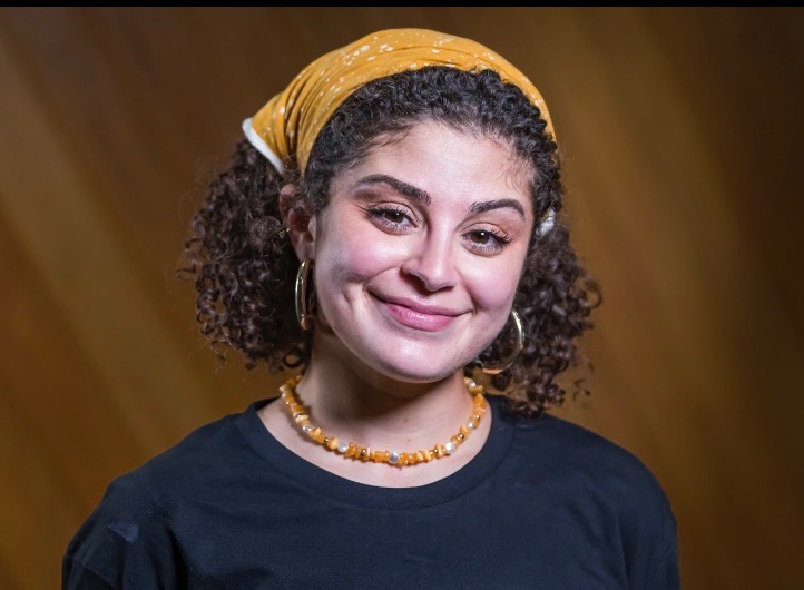 Mona Abhari, young Palestinian woman with dark curly hair held back by a yellow bandana, wearing gold hoop earrings, soft pink makeup, an orange shell necklace, and a black shirt. She is smiling with a slight head tilt towards the camera.