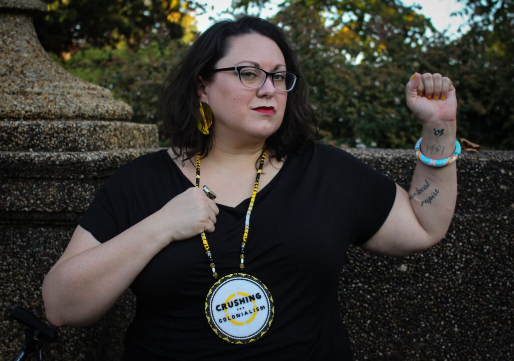 Jen Deerinwater, a white coded femme presenting person with black glasses, brown, wavy, shoulder-length hair worn down, sits in a park with stone masonry and trees behind hir. Jen’s left fist is raised in the air, exposing tattoos and a Native designed bangle bracelet. Hir right hand is holding onto one side of a Native made beaded medallion with the Crushing Colonialism logo in the center. Jen is wearing a black, short sleeve shirt and large, yellow earrings.