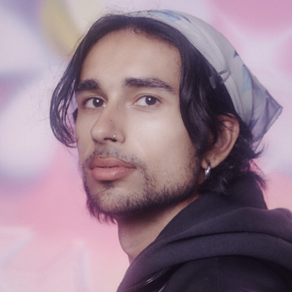 A headshot of Noah Gokul, a Queer person of color, against a soft floral pink background. They facing the camera, wearing a light-colored headscarf and black hoodie.