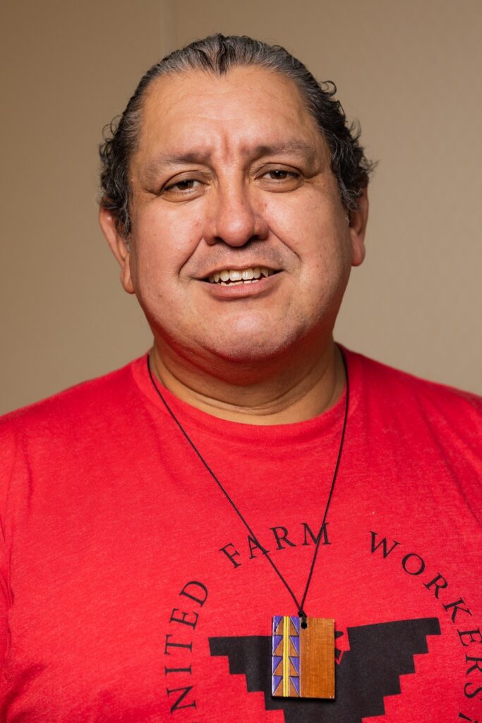 Héctor has long hair pulled back in a ponytail, and their black hair now has streaks of gray. Héctor is wearing a red shirt that reads "United Farm Workers" and has a redwood pendant tied around their neck with a string.