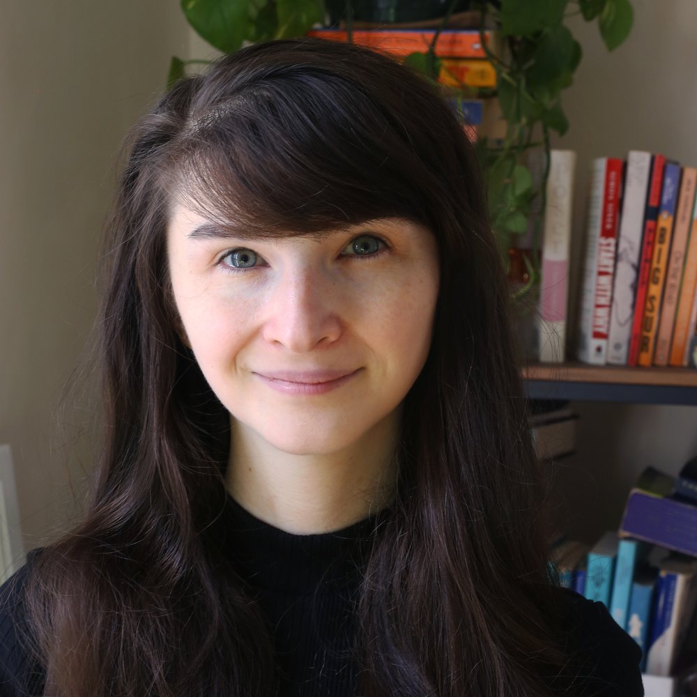Jessie Roth is a woman with shoulder-length dark brown hair wearing a black shirt. She is in front of a color-coded bookshelf topped by a sprawling green plant. She is smiling at the camera.