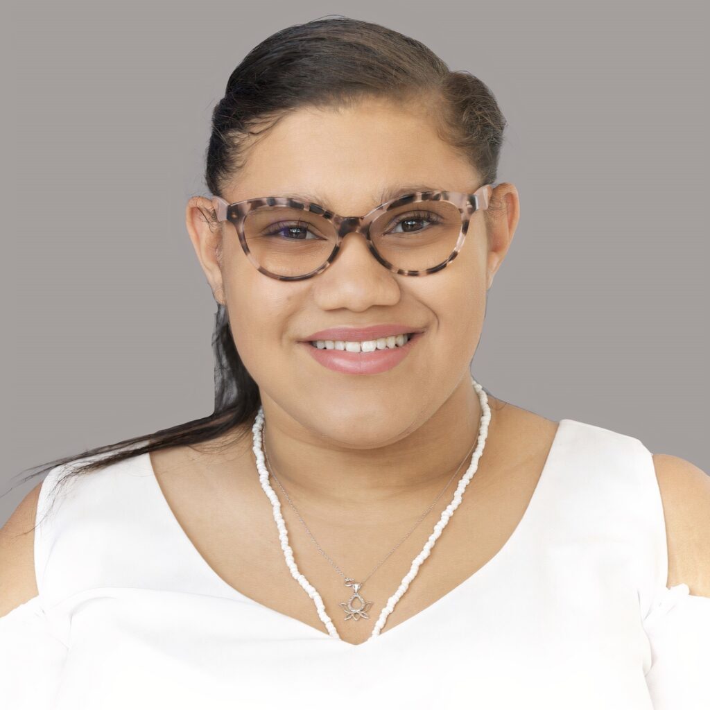 An image of Daphne Frias, a young disabled Latina with brown shoulder-length hair, wearing brown patterned glasses, a white necklace, and a white off-the-shoulder blouse. She is smiling at the camera.