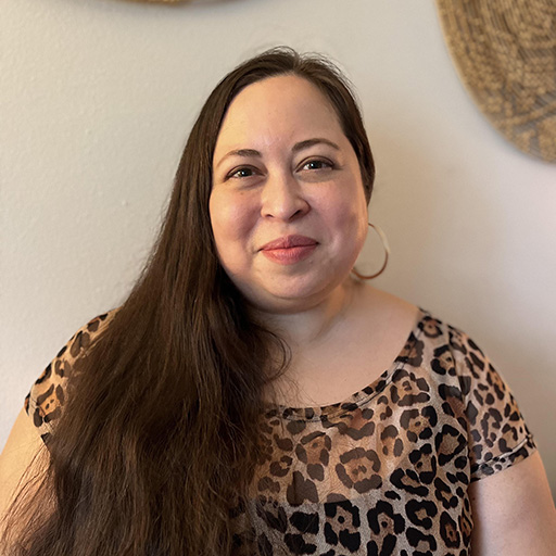 A fat light-skinned latine-indigenous person wearing a leopard print dress smiles directly at the camera. They are wearing gold hoop earrings with brown waist-length hair pulled to the front and lying over their left side shoulder. Behind them is a mostly off-white wall with some tan circular basket-woven style art on the wall. 