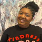 Razz is a Black woman with brown skin and long black marley twists pulled into a big bun on the top of her head. She's standing in front of a blue and white abstract painting of a tree wearing a black sweater with the word "kindness" stacked in red and pink.