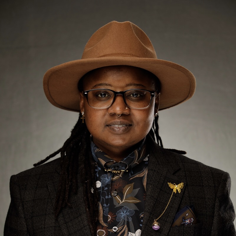 Shaena Johnson, a young Black woman with medium length, black locs, wearing a tan hat, black, brown and burgundy floral shirt, brown plaid jacket, eye glasses.  She is smiling partially, while looking at the camera.