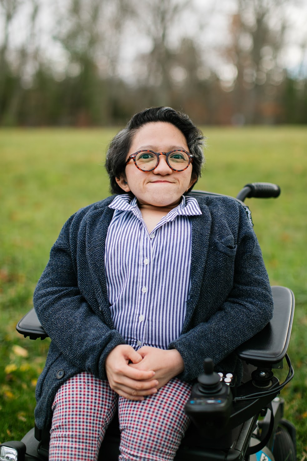 Sandy Ho, an Asian American woman in a power wheelchair, sits in a field of grass looking confidently into the camera. She is wearing round glasses, a gray sweater over a blue striped collar shirt, and red checkered pants.