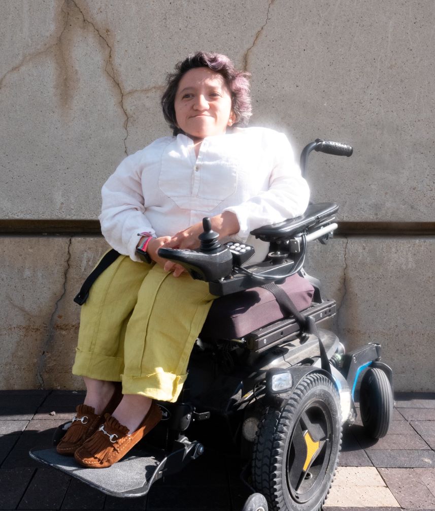Photo taken by Adrian O. Walker. Sandy Ho is sitting outside against a wall in a power wheelchair. She’s wearing a long sleeved white shirt, yellow pants, and brown shoes with a silver buckle. She has short dark wavy hair, and she is smiling at the camera.