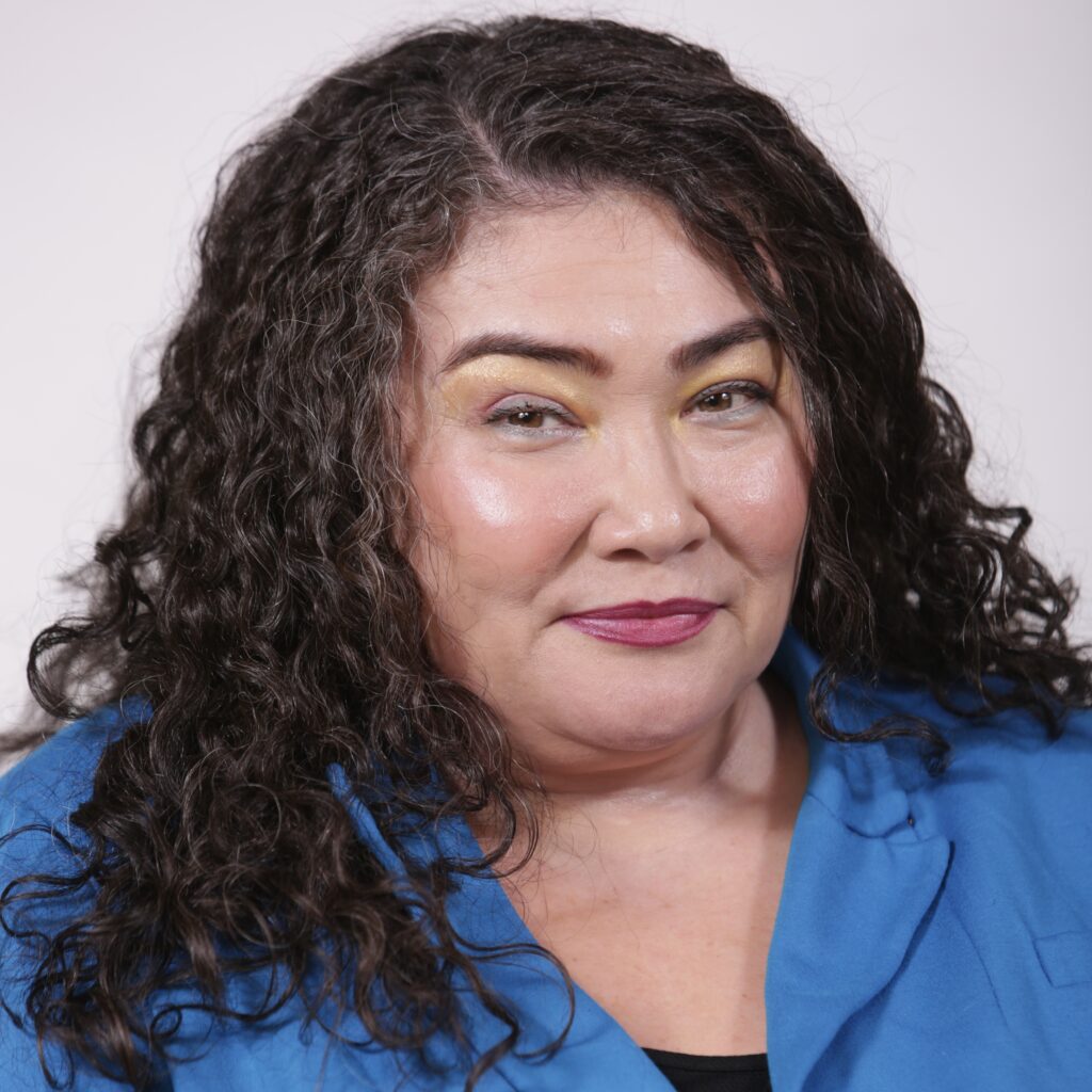 Rahnee Patrick is an Asian American person against a white background. She has curly, dark, gray-streaked shoulder-length hair. They are wearing a bright blue blazer with a black top and looking confidently into the camera.