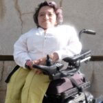 Photo taken by Adrian O. Walker. Sandy Ho is sitting outside against a wall in a power wheelchair. She’s wearing a long sleeved white shirt, yellow pants, and brown shoes with a silver buckle. She has short dark wavy hair, and she is smiling at the camera.