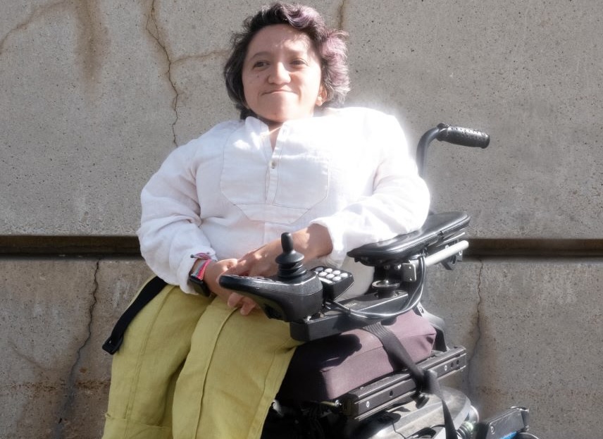 Photo taken by Adrian O. Walker. Sandy Ho is sitting outside against a wall in a power wheelchair. She’s wearing a long sleeved white shirt, yellow pants, and brown shoes with a silver buckle. She has short dark wavy hair, and she is smiling at the camera. 