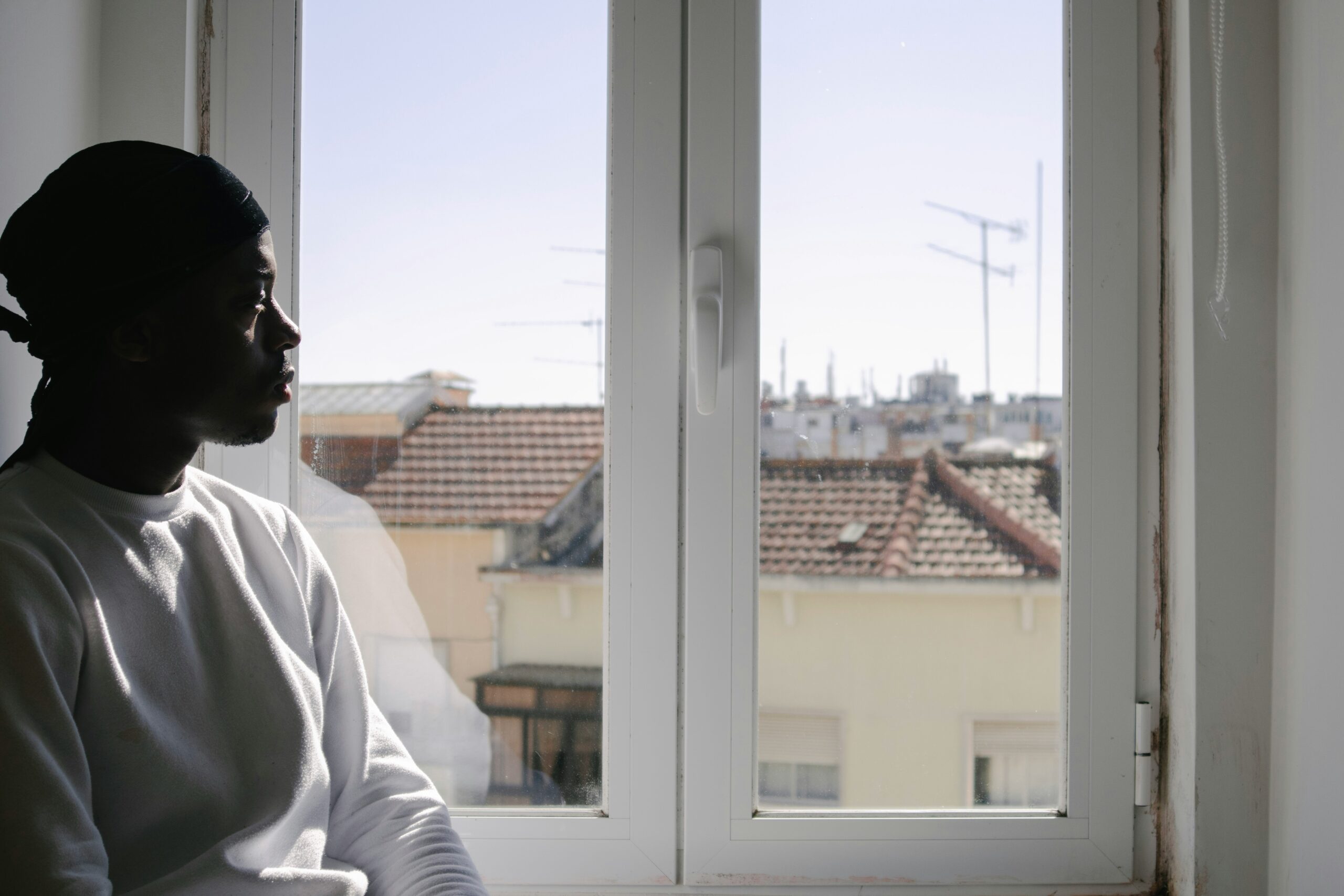 A side profile shot of a Black man gazing solemnly out of a window on a sunny day. The man wears a white sweatshirt and a black durag. Through the window are nearby apartments and a blurry city skyline.