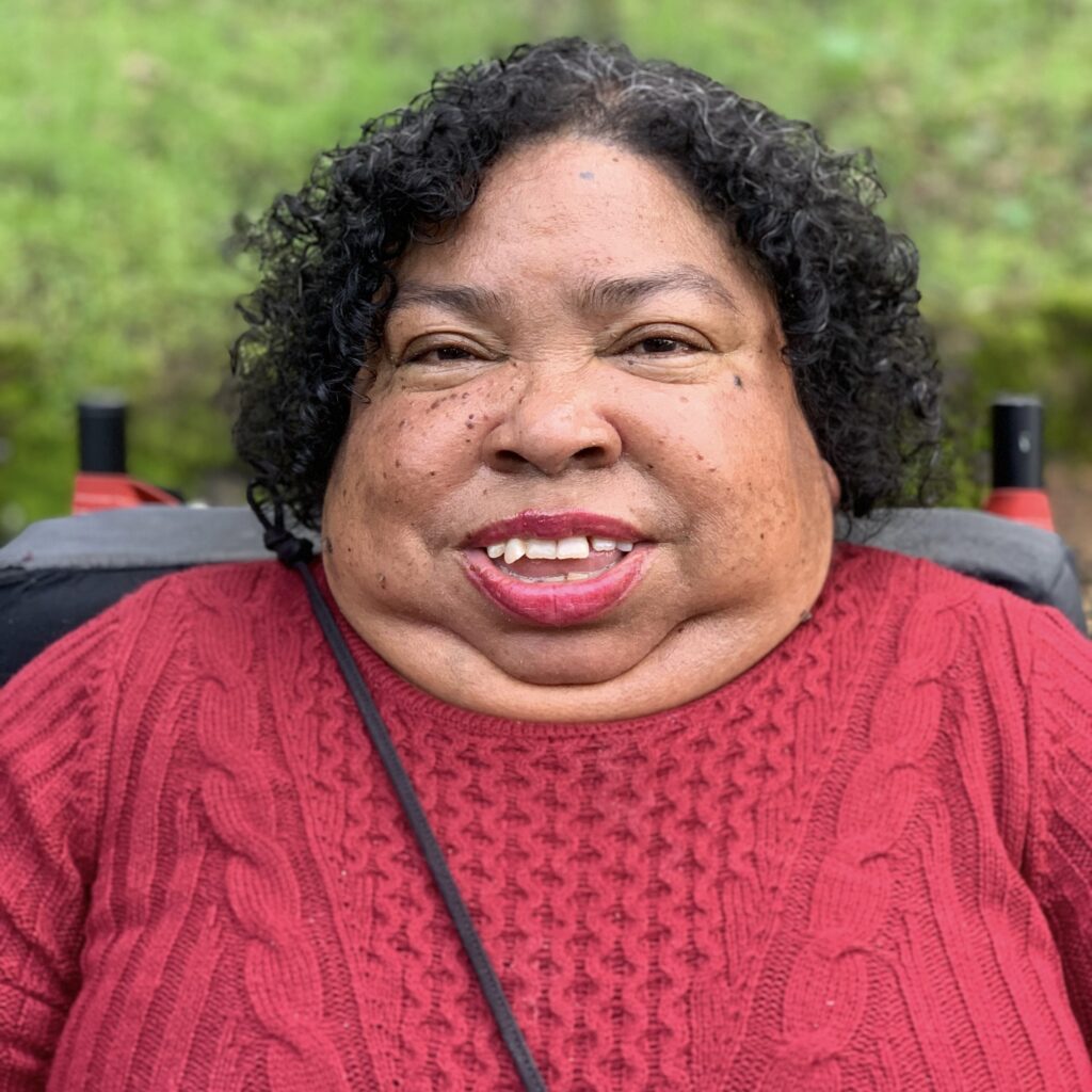 Nikki Brown-Booker is a Filipino/Black woman with short dark curly hair, wearing a red sweater. Sitting in her wheelchair, she smiles at the camera with a blurred greenery exterior in the background.