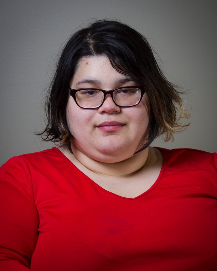 Fat light skinned Latina woman with chin length hair, who is wearing purple glasses and a red dress while smiling at the camera.