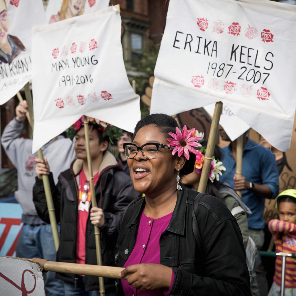 A picture of Mel Brown with people in the background holding signs. Mel is a Black person holding a sign and wearing a pink flower in her hair, black rimmed glasses, and a black jacket over a pink top.