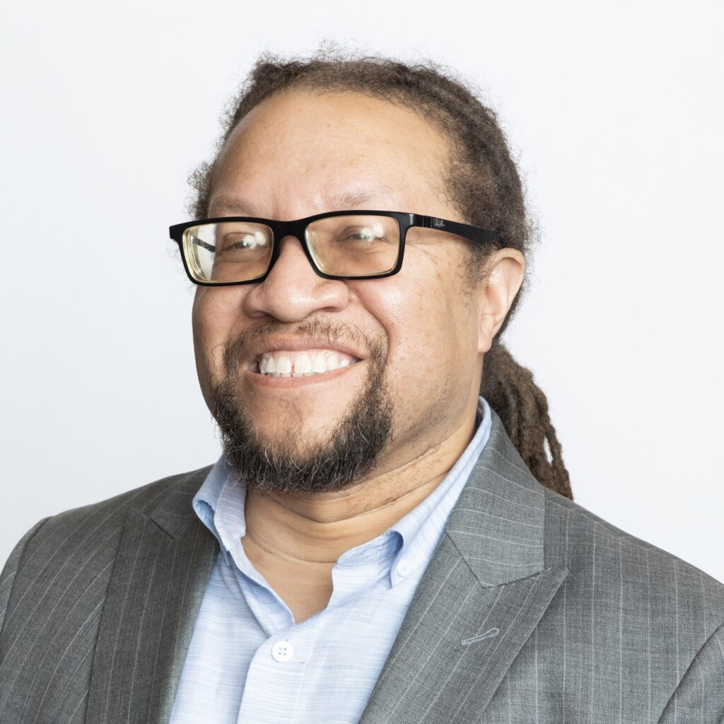 A headshot of Patrick Cokley, a light skinned African American man with dreadlocks pulled back, wearing dark glasses and a light gray suit. He is smiling at the camera.