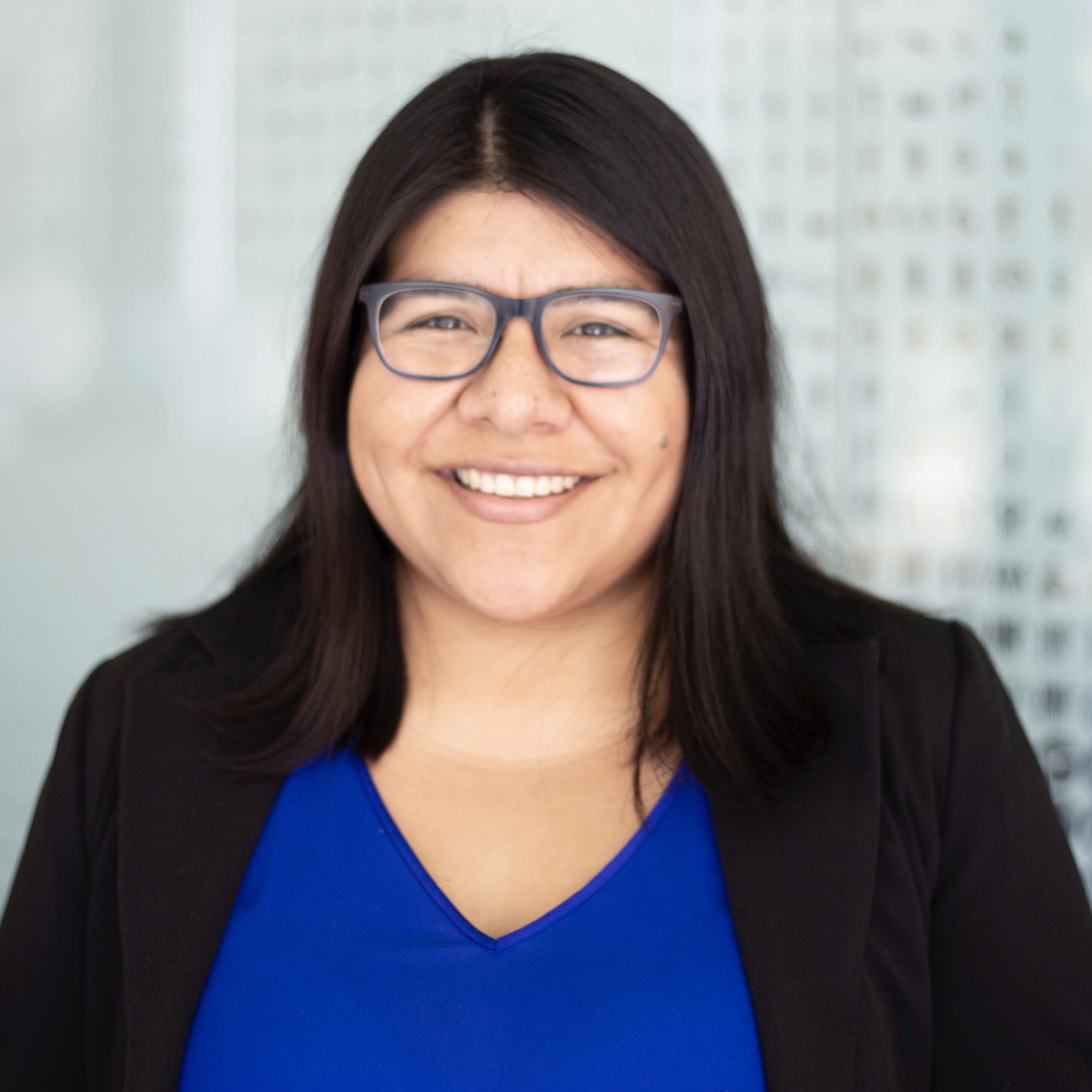 Latina woman with shoulder length dark brown hair, dark brown eyes with black rimmed glasses and a big smile wearing a cobalt blue blouse and black blazer. She is standing in front of a light aqua glass door.