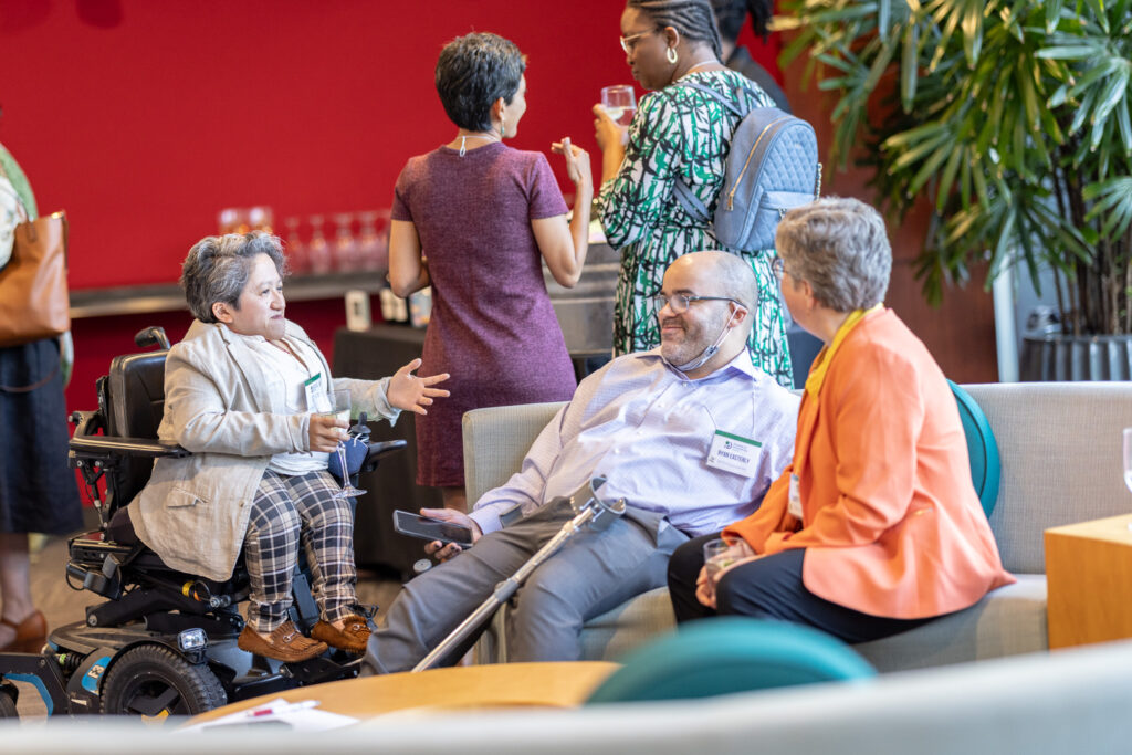 A candid photo of Sandy Ho, Ryan Easterly, and Kristy Trautmann engaged in conversation during the 2024 Meeting of the Presidents’ Council of the Disability & Philanthropy Forum. Sandy is in her power chair, and Ryan and Kristy are seated on a gray sofa. Behind them are are bright red wall and other attendees chatting with their backs to the camera.