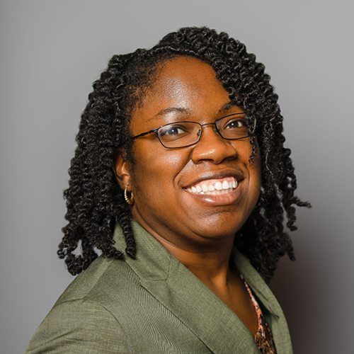 Britney Wilson is a Black woman with black coily hair and dark-rimmed glasses. She is smiling at the camera against a gray background, wearing small hoop earrings and an olive green blazer.