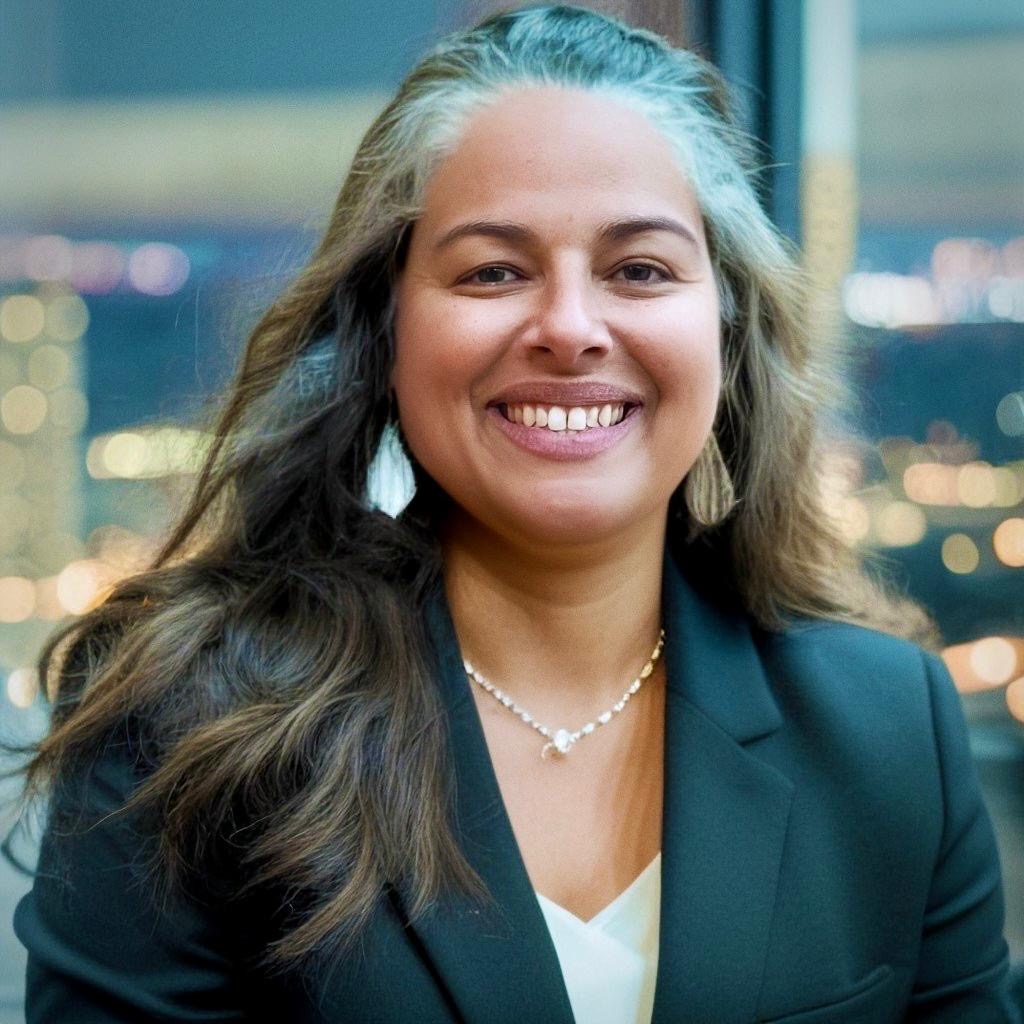 A Latina woman with long hair and gray streaks smiles warmly. She wears a black blazer, white top, and a necklace.