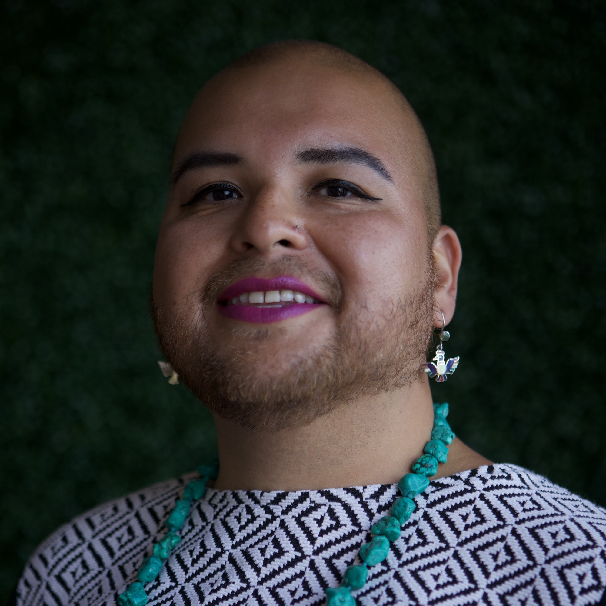 Aldita Amaru Gallardo, a disabled Indigenous queer trans woman with low shaved hair, black eyeliner, and a close-shaven beard. She is smiling at the camera against a blurred dark green background, wearing a black and white patterned top, aqua blue necklace, colorful earrings, and pink lipstick.