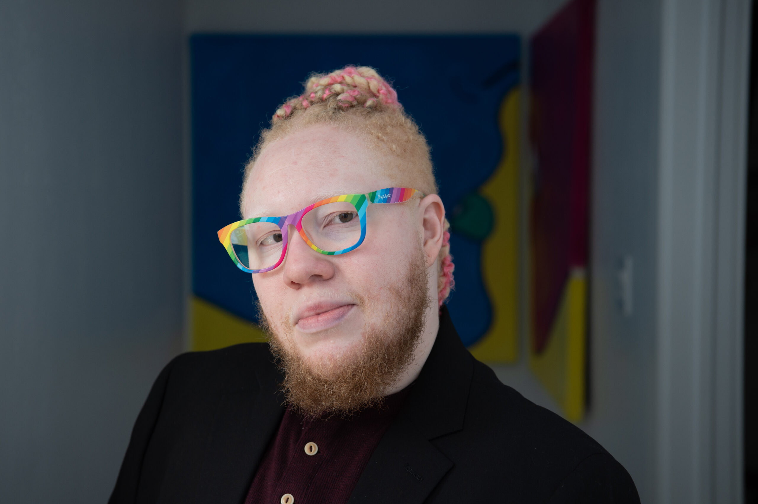 An albino Black trans man with pink and blond hair wearing rainbow glasses, a maroon shirt and black blazer looks toward the camera in a white hallway with two colorful paintings.