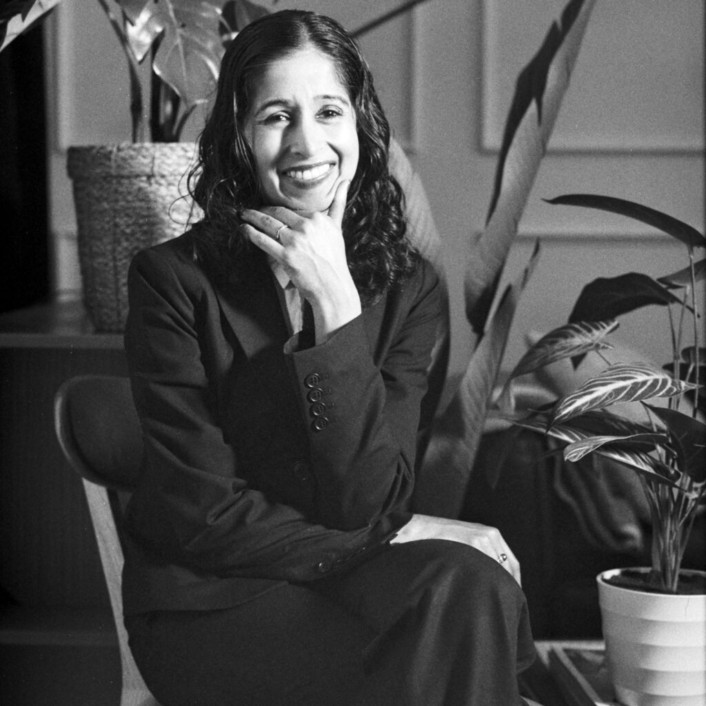 Qudsiya Naqui, a South Asian woman with brown skin, shoulder-length curly dark hair, and dark eyes, sits in a chair with her legs crossed, smiling with her chin resting on one hand. Though the image is in black and white, she sports a dark suit with a red tie, and is surrounded by ferns.
