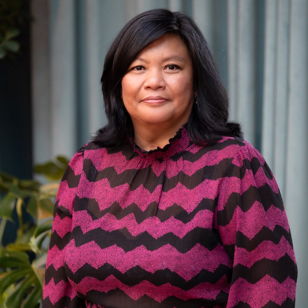 Marissa Tirona, a mid-life Filipina with shoulder-length, wavy Black hair, wearing a black and magenta striped dress. She is smiling calmly at the camera.