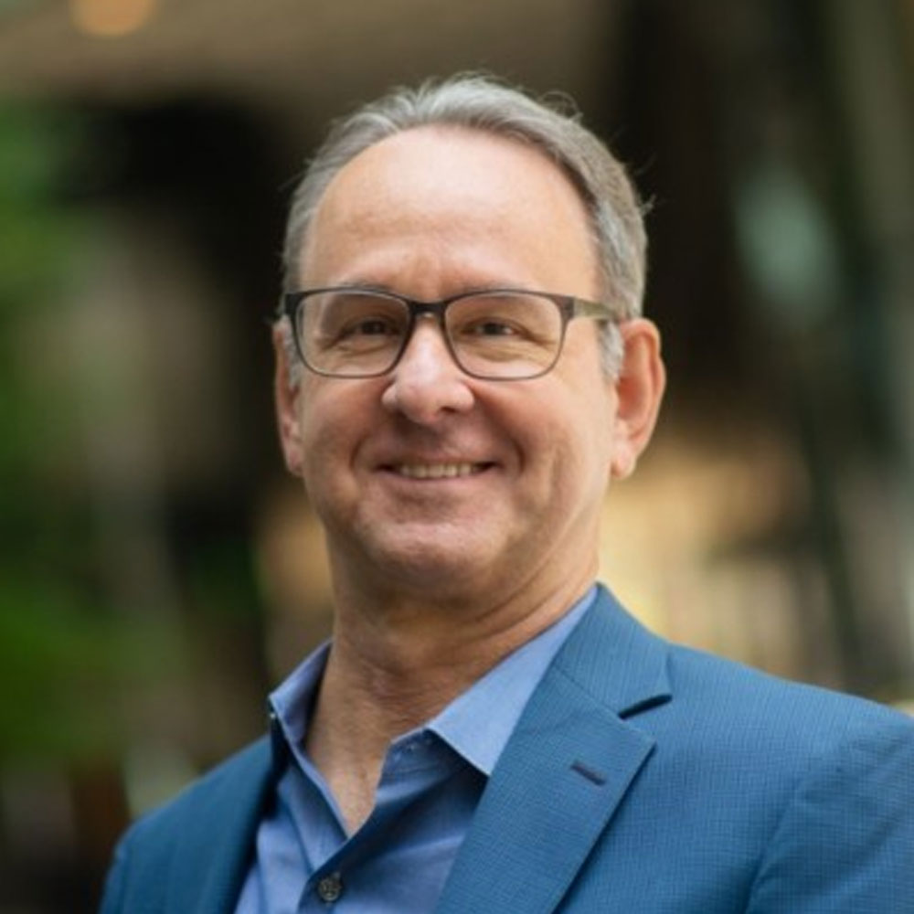 Henry Claypool is a white male with brown and grey hair wearing glasses, a blue button-down shirt with a blue blazer.  He is smiling at the camera.
