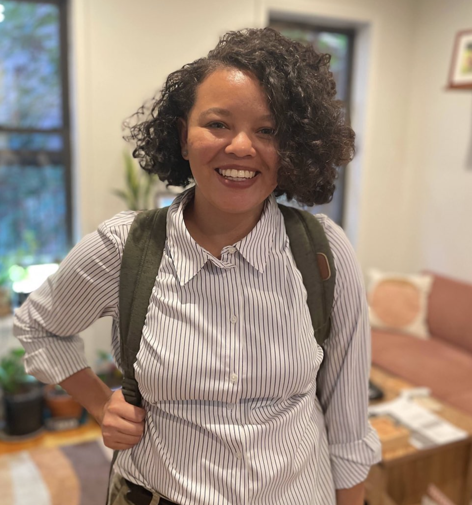Jalyn is a Black, Japanese person with short, dark brown curly hair. They stand indoors smiling while wearing a white button-up shirt with black stripes and an olive green backpack. In the background, there are windows, plants, and modern pink furniture.