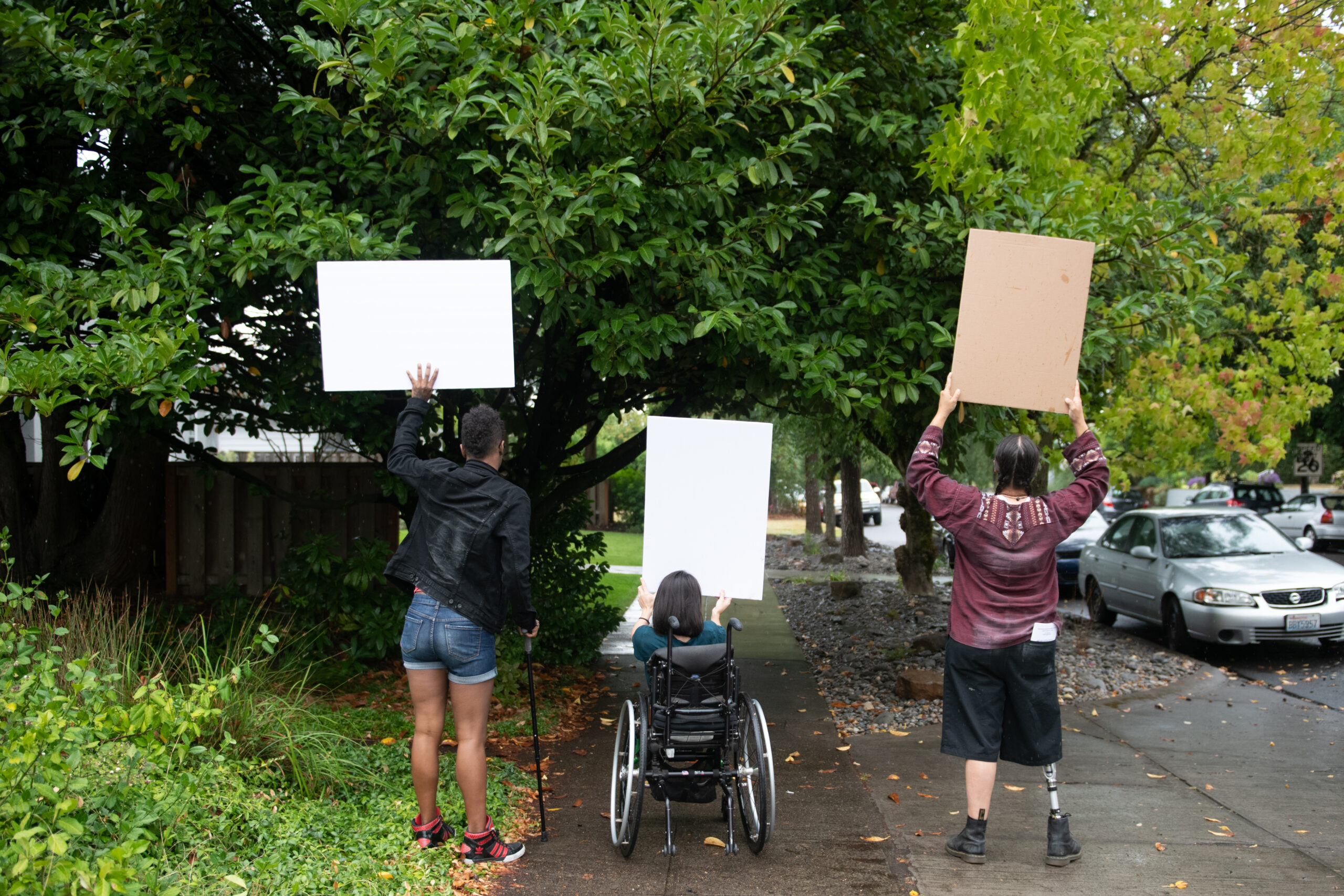 Three disabled people of color (a Black non-binary person with a cane, a South Asian person in a wheelchair, and an Indigenous Two-Spirit person with a prosthetic leg) block a neighborhood street while holding up cardboard signs. The photo by Chona Kruger is shot from behind everyone. Photo courtesy of Disabled and Here.