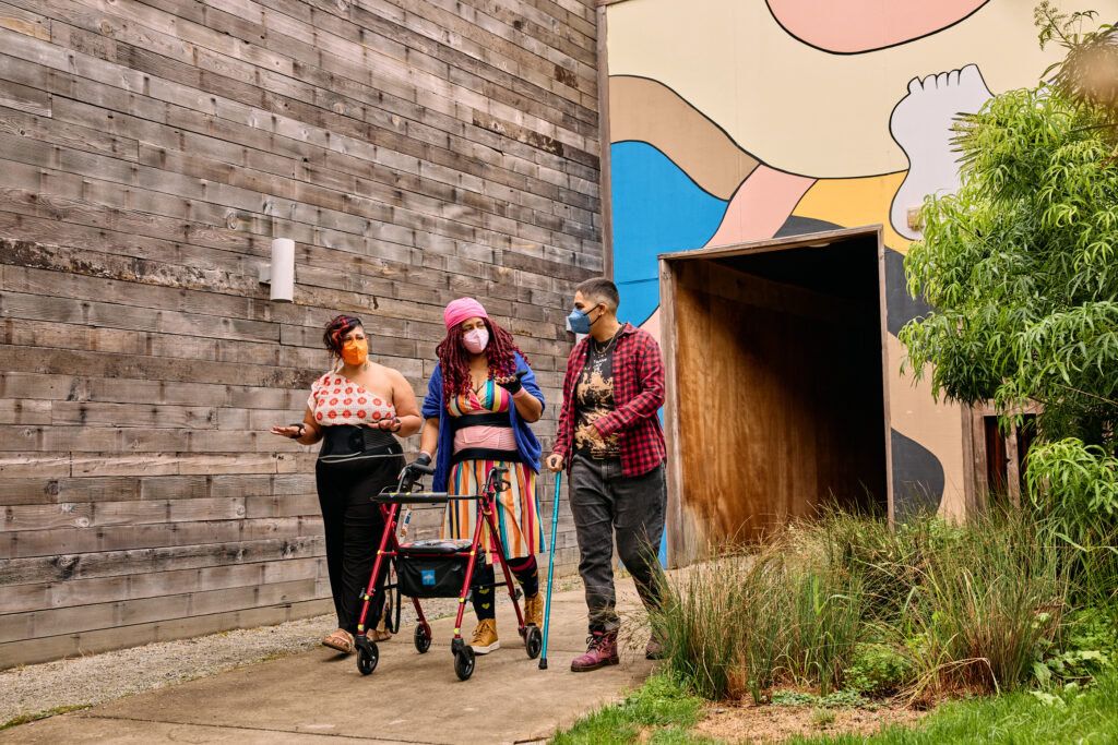Three masked disabled people of color chat while strolling through an outdoor courtyard. On the left, an Indigenous woman wears glasses, wrist support braces, and waist trainers paired with a off-shoulder top and wide leg pants. In the middle, a Black non-binary woman walks with a rollator and wears waist trainers with ice pack inserts over their rainbow striped dress. On the right, a Latinx non-binary person walks while using a teal functional grip cane.